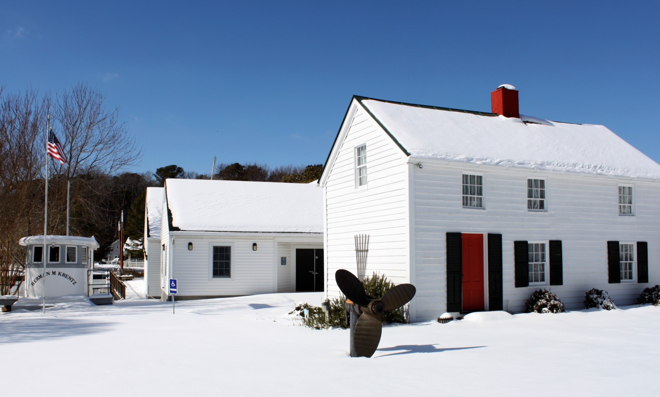 A two story white clapboard house with dark shutters and a chimney and a one story white out building behind it