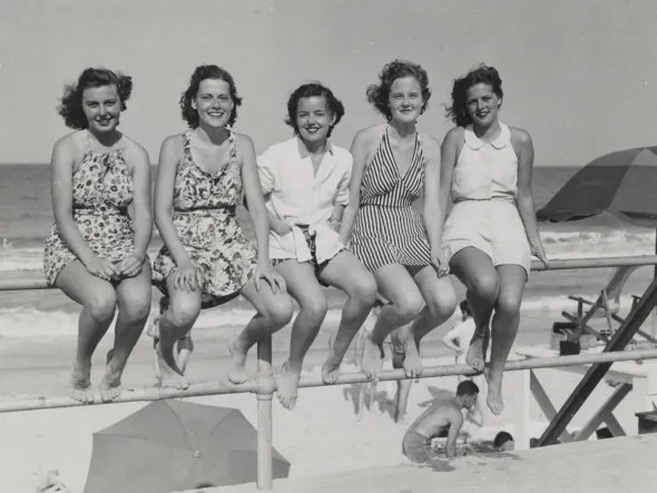 A row of women in 1950s-era dresses and bathing suits sit on a metal railing in front of an ocean beach.