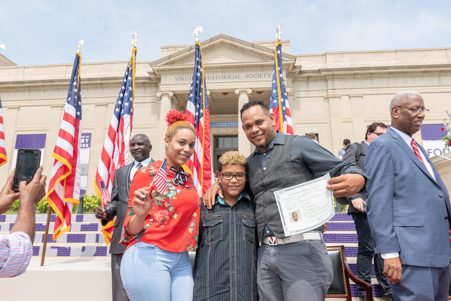 A mom, dad, and son pose on the steps of the VMHC in front of US flags holding a certificate 
