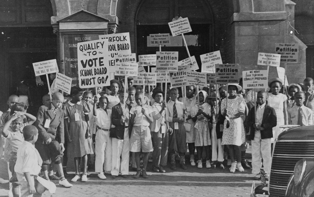  Demonstrators at a Norfolk parade holding signs that read "WE WANT OUR TEACHERS EQUALLY PREPARED AND EQUALLY PAID” and “QUALIFY TO VOTE SCHOOL BOARD MUST GO,” as they protest inequality between black and white schools in 1939. 