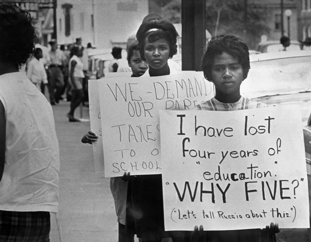 Black and white photograph of female student in the forefront of a protest in Prince Edward County, 1963, holding a sign that reads, “I have lost four years of “education.” “WHY FIVE?” (“Let’s tell Russia about this.”)