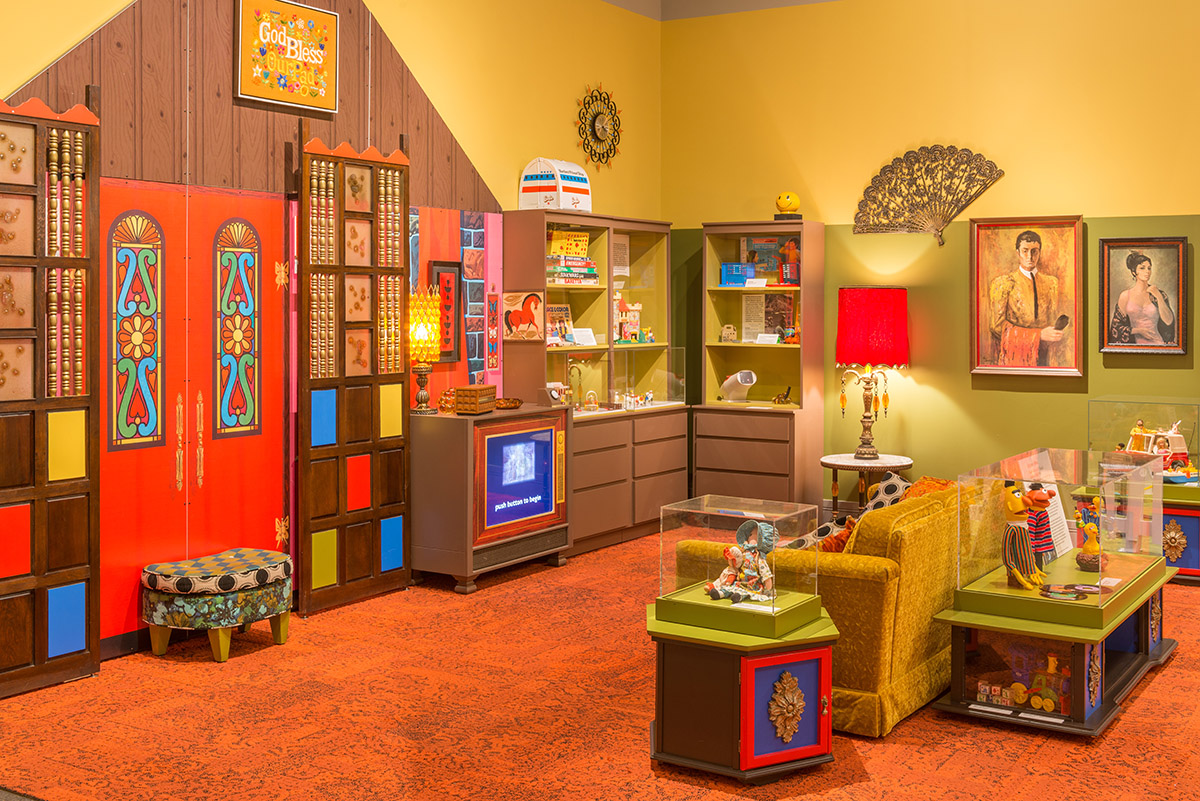 An exhibition display designed like a 1970s living room with couch, side tables, tv, wall art, and rug from the decade