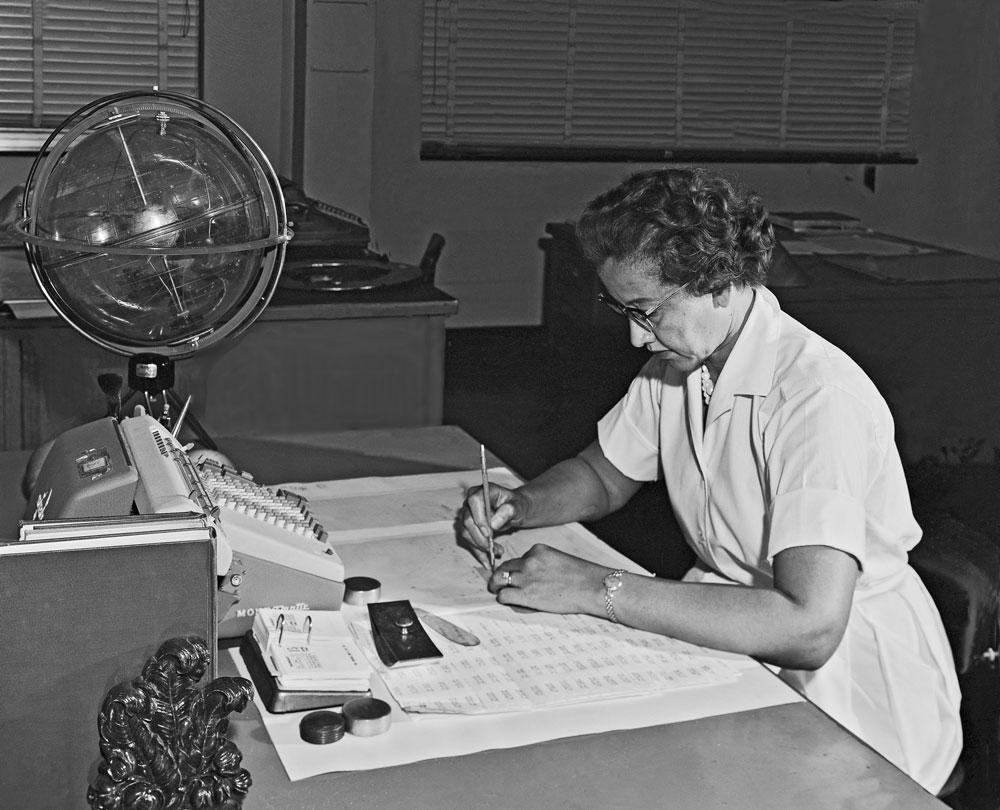 Black and white photo of Katherine Johnson sitting at a desk writing on a large printed spreadsheet. A globe and machine sit on the desk in front of her.