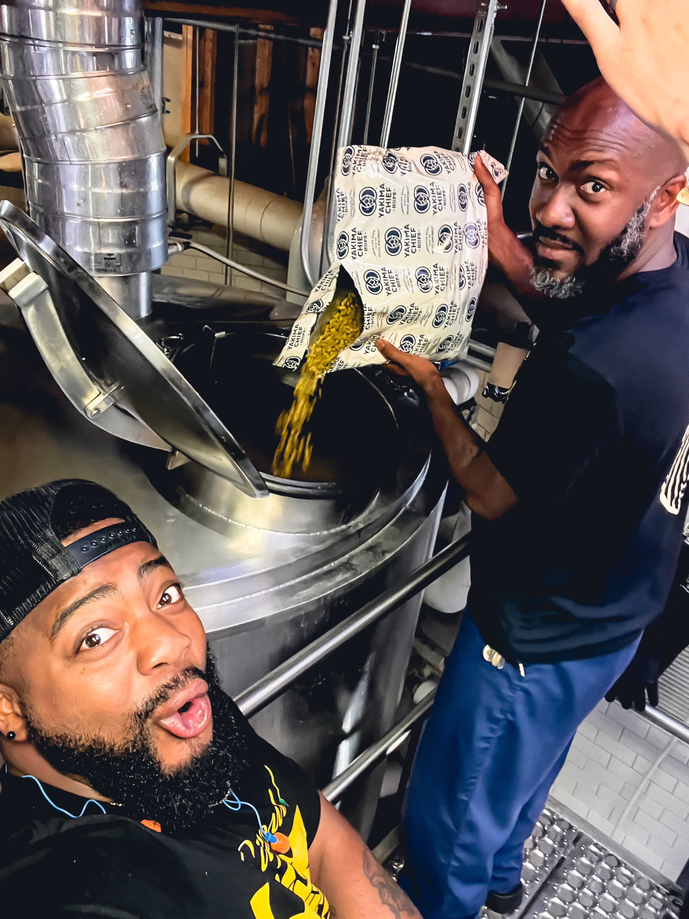 Two Black men add hops to a stainless steel barrel in the beer brewing process