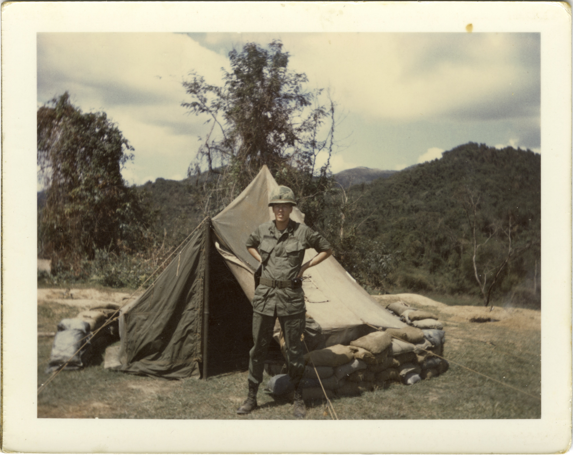 A soldier stands in front of tent in a mountainous landscape.