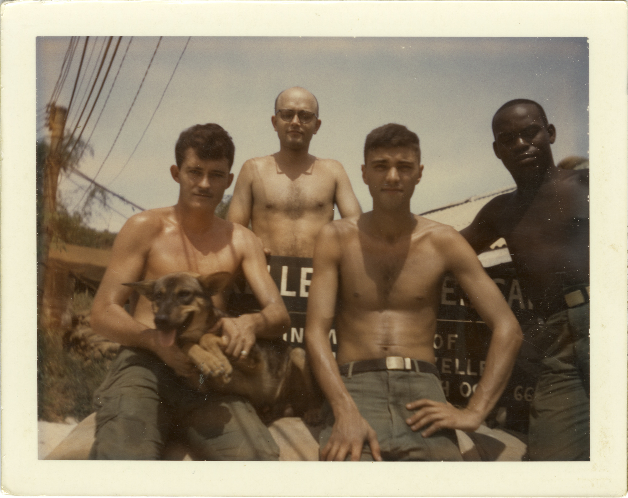 Four soldiers without shirts and wearing military green fatigue pants sit holding a small dog