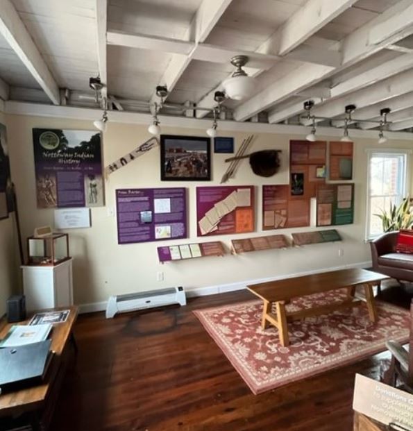 An interior room with wooden floors and beamed ceiling with a rug and wooden table and interpretive panels along the wall