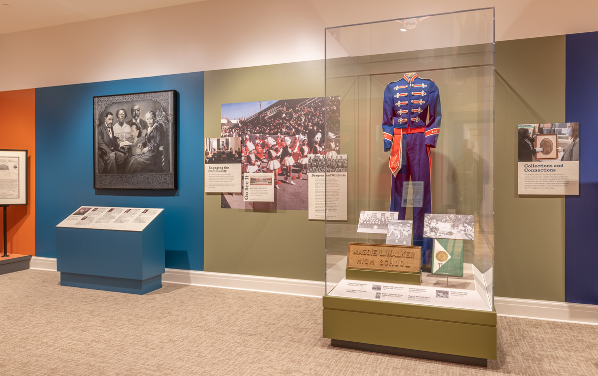 A photo of an exhibition with a painting and photographs on the gallery walls, and a marching band uniform in a display case