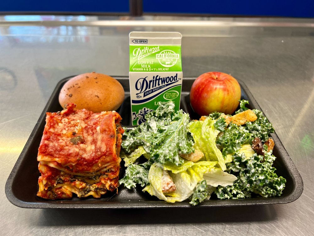 A school lunch tray with roll, milk, apple, kale salad, and veggie lasagna