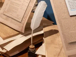 Manuscripts and a feather pen on a table with an exhibit label showing a Twitter post