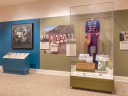 A photo of an exhibition with a painting and photographs on the gallery walls, and a marching band uniform in a display case