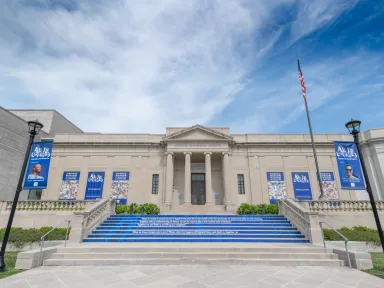 A photo of the front of the museum shows blue artwork on the stairs and blue bannerw with the faces of artists and colorful illustrations.