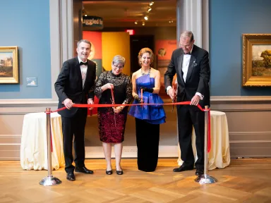 Four people stand in a wood-floored room cutting a red ribbon in front of a new gallery exhibition 
