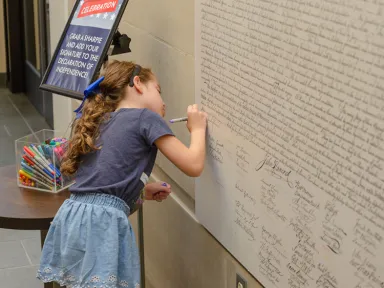 A young child signs a large replica of the Declaration of Independence