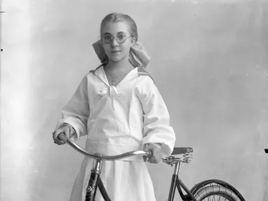 Black & white photo of a young girl with a ponytail and glasses with a bike beside her