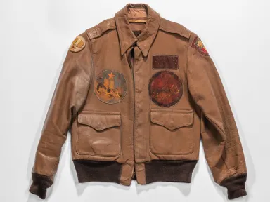 A brown leather jacket with sewn patches on the front and a flapped pocket on the bottom left and right sides