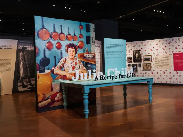 The exhibition entrance for Julia Child: A Recipe for Life features wood bloors, a large blue dining table, and an oversized photo of Julia in her kitchen in front of a wall of copper pots.