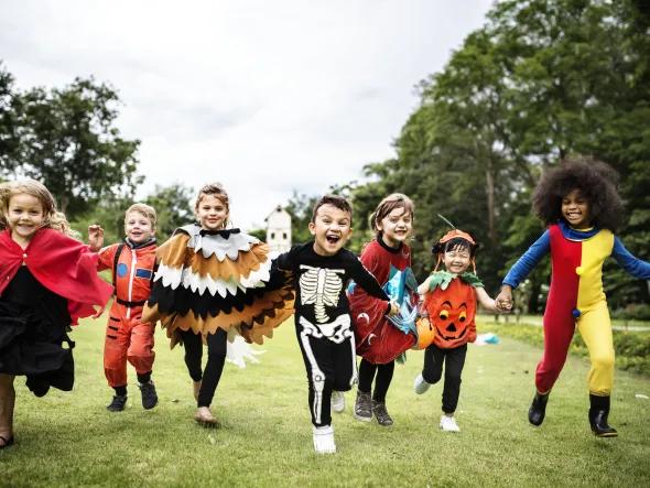 A group of children in Halloween costumes run across a grassy field with trees in the background