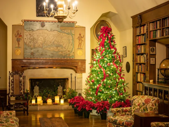 A Tudor-style library room features a Christmas tree decorated with lights and red ribbons, with poinsettias beneath it, wide fireplace filled with candles, globe, bookshelves, 