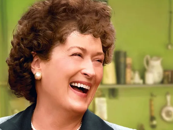 Meryl Streep dressed as Julia Child laughs in a green kitchen.