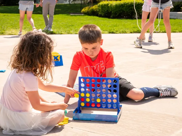 Little girl and boy play ConnectFour on a patio with green lawn and children playing behind them.