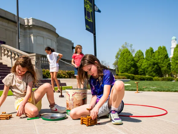 Girls playing with Lincoln Logs and HoolaHoops on a patio with green grass behind them.