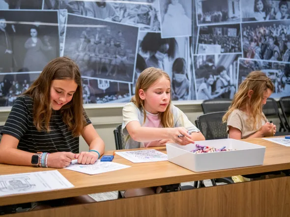 Three girls sit at a table working on crafts with a mural of historical black and white photos behind them.