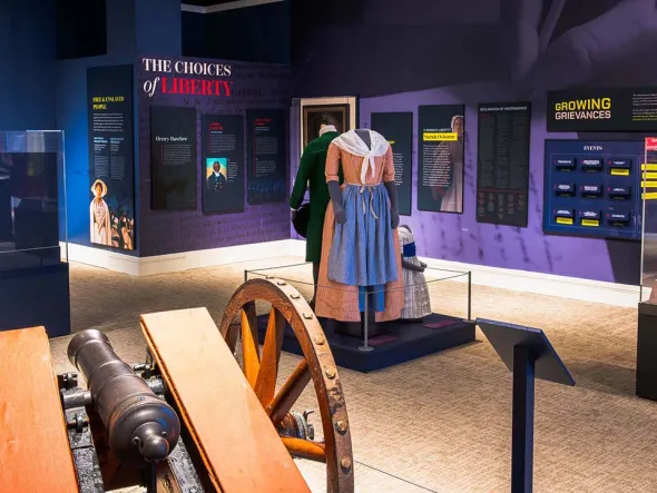 A museum exhibit featuring historical displays, period clothing on mannequins, and an antique cannon in the foreground. Informational panels on the walls provide context about historical figures and events, with a section labeled "Growing Grievances" visible in the background.