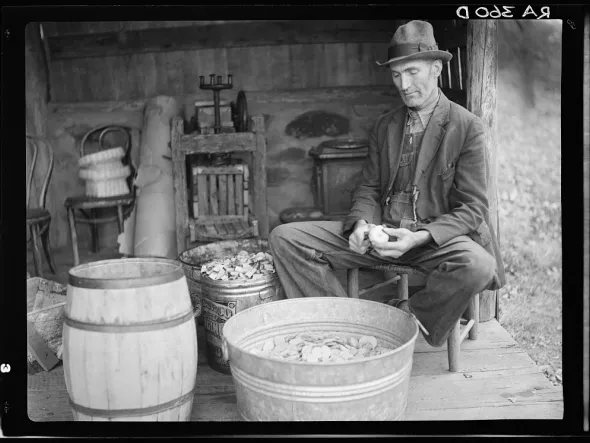 A black and white photo of a man in long jacket, pants, and domed hat, sitting in front of a barrel and washtub