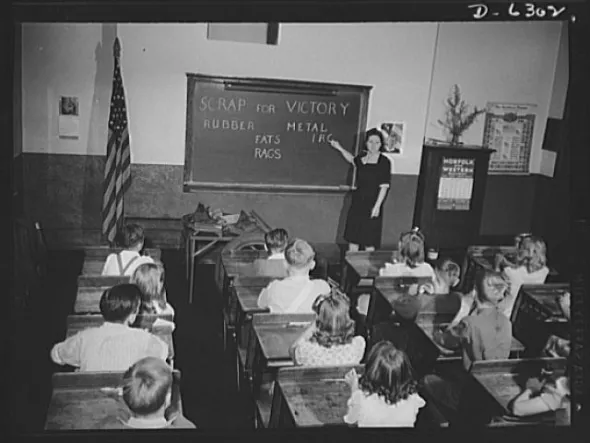 A black and white photo of a classroom. The words SCRAP FOR VICTORY, RUBBER, METAL, FATS, RAGS are written on the chalkboard.