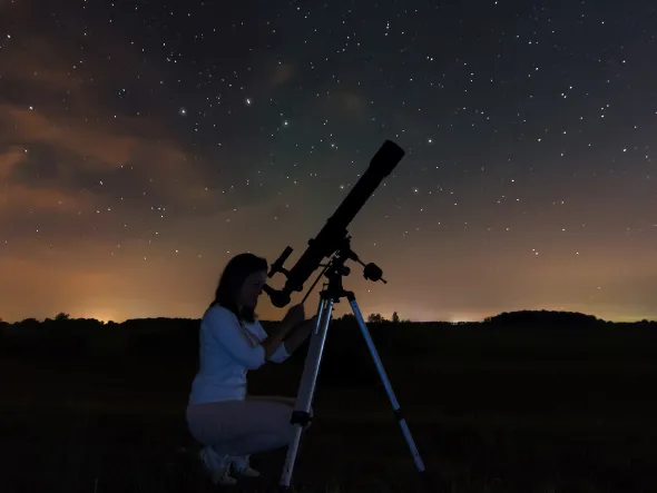 A woman looks through a telescope with a starry sky behind her