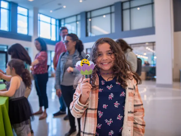 A child smiles while holding a paper craft of a bee in a flower
