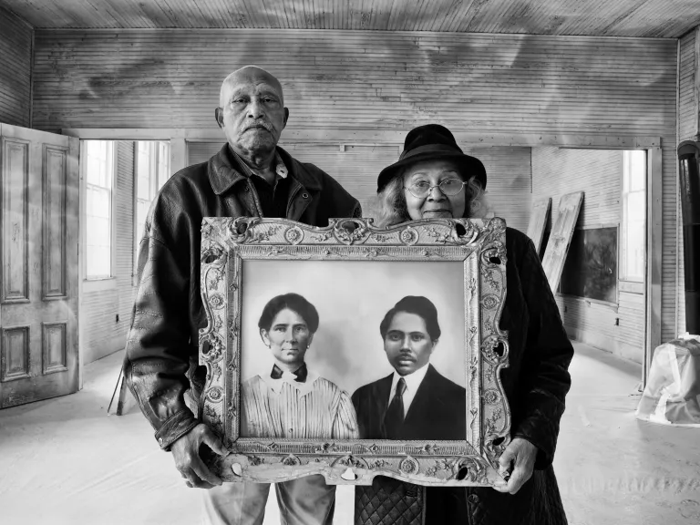 Two people hold an ornately frame painted portrait of two people in early 1900s dress