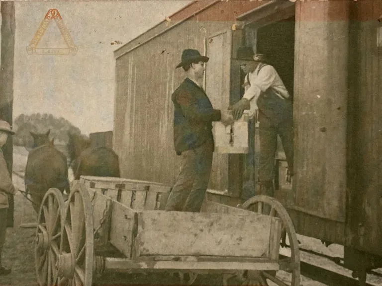 A black and white photo of a man in a horse-led cart handing a crate to a  man on a train car