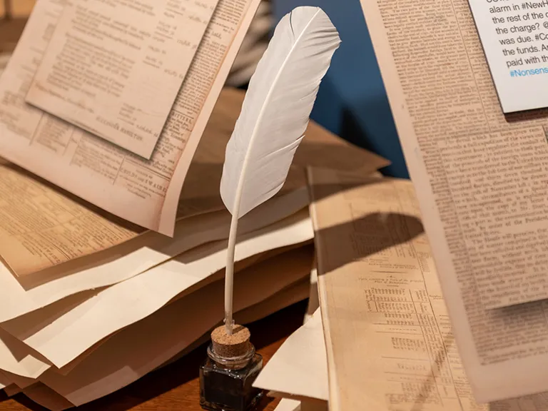 Exhibition display from Founding Frenemies Manuscripts and a feather pen on a table with an exhibit label showing a Twitter post