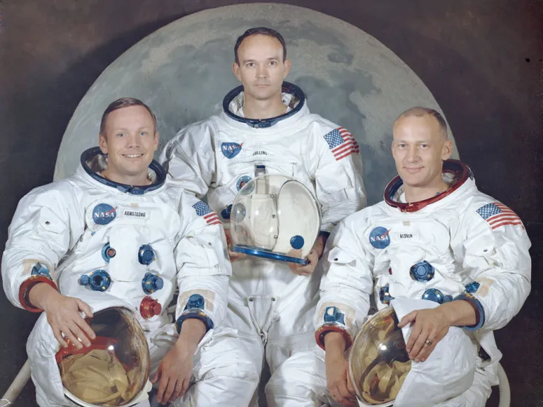 The crew of Apollo 11: 3 astronauts in full space suits hold space helmets in front of a backdrop depicting the moon