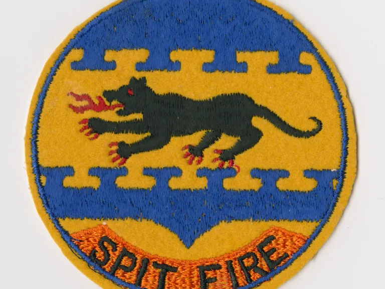 Embroidered felt patch, "SPIT FIRE." Circular patch features a black, fire-breathing panther in the center embroidered on a yellow felt field. Blue emboidery is above and below the panther with "SPIT FIRE" embroidered in black on an orange embroidered field at the bottom.