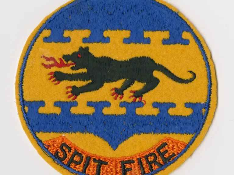 Embroidered felt patch, "SPIT FIRE." Embroidered felt patch, "SPIT FIRE." Circular patch features a black, fire-breathing panther in the center embroidered on a yellow felt field. Blue emboidery is above and below the panther with "SPIT FIRE" embroidered in black on an orange embroidered field at the bottom.