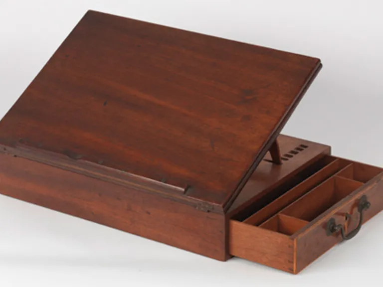 A rectangular wooden desk, with writing surface propped at an angle and drawer opened