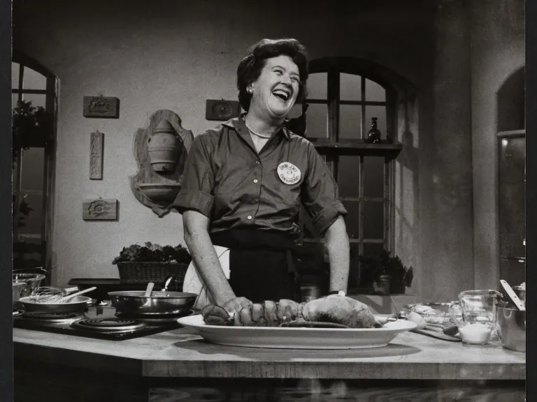 A black and white photo of Julia Child in her kitchen standing behind a countertop filled with cooking implements and ingredients