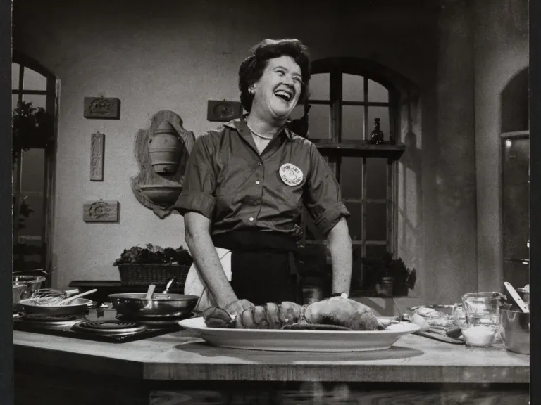 Julia Child – Photography by Paul Child © The Schlesinger Library, Radcliffe Institute, Harvard University A black and white photo of Julia Child in her kitchen standing behind a countertop filled with cooking implements and ingredients