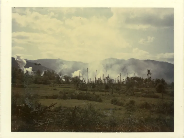 Photos of a green field with trees, smoke in the distance, and mountains beyond