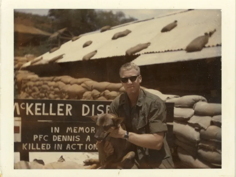 A man in military uniform with a dog outside of a low barracks building