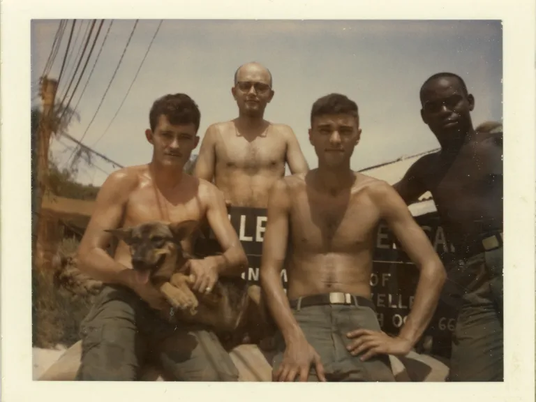 The dog “Keats” and several of Lt. Rasmussen’s men. Four soldiers without shirts and wearing military green fatigue pants sit holding a small dog