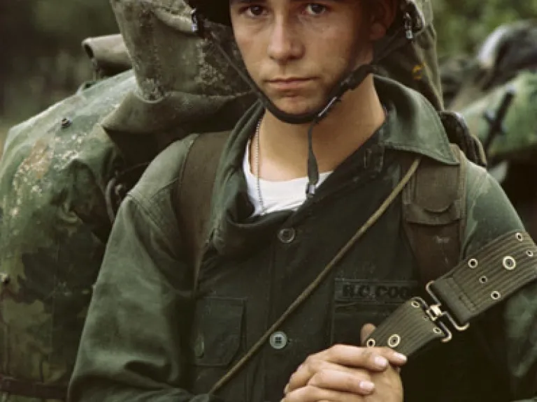 A young solider in military fatigues and helmet carrying a camo backpack