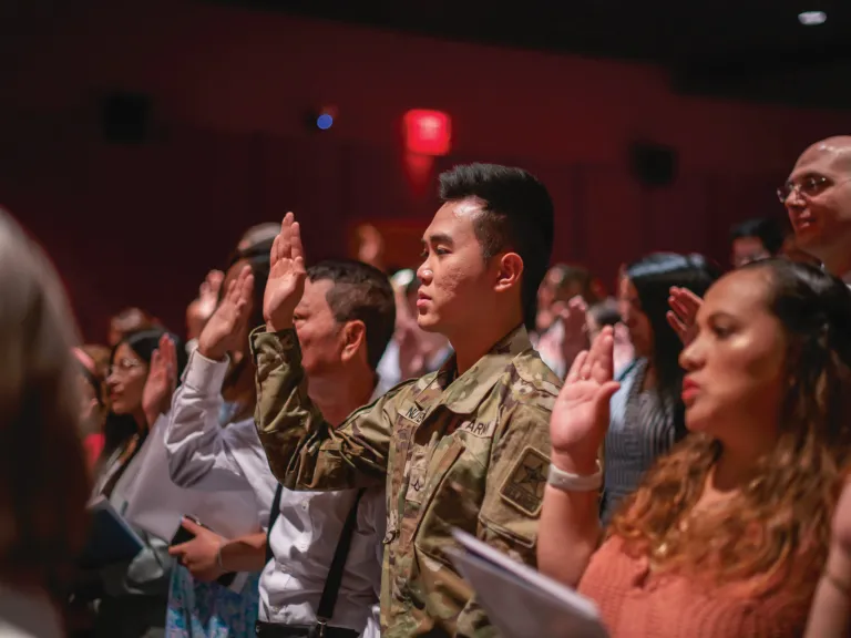 People stand in an auditorium with their right hands raised, taking the U.S. Oath of Citizenship