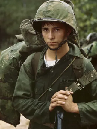 A young solider in military fatigues and helmet carrying a camo backpack