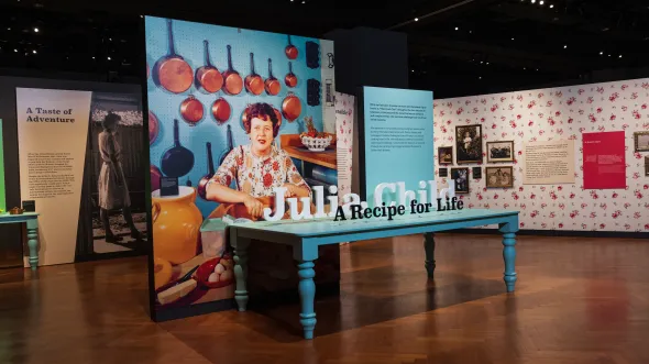 The exhibition entrance for Julia Child: A Recipe for Life features wood bloors, a large blue dining table, and an oversized photo of Julia in her kitchen in front of a wall of copper pots.