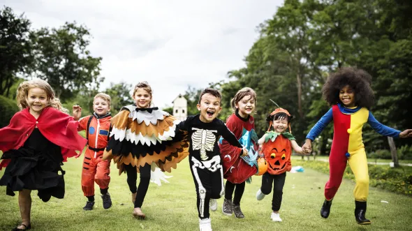 A group of children in Halloween costumes run across a grassy field with trees in the background