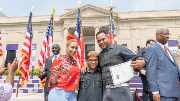 A smiling family stand in front of American flags holding a Citizenship certificate